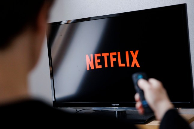 Logo du service de streaming Netflix visible sur un écran de télévision avec un spectateur masculin vu de derrière tenant sa télécommande vers l'écran