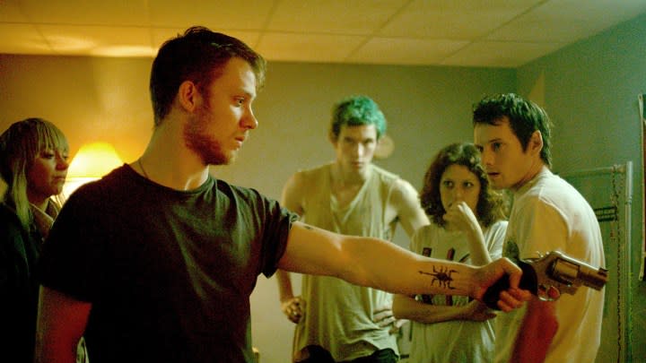 Plusieurs personnes dans une pièce, l'une tenant une arme pointée vers la droite dans Green Room.