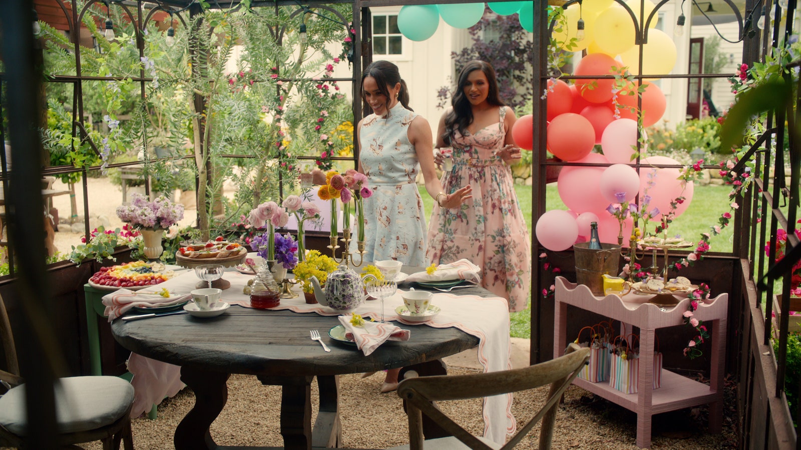 Meghan Markle dans une structure extérieure entourée de fenêtres, dans un jardin avec des ballons Mindy Kaling et une table dressée pour dîner
