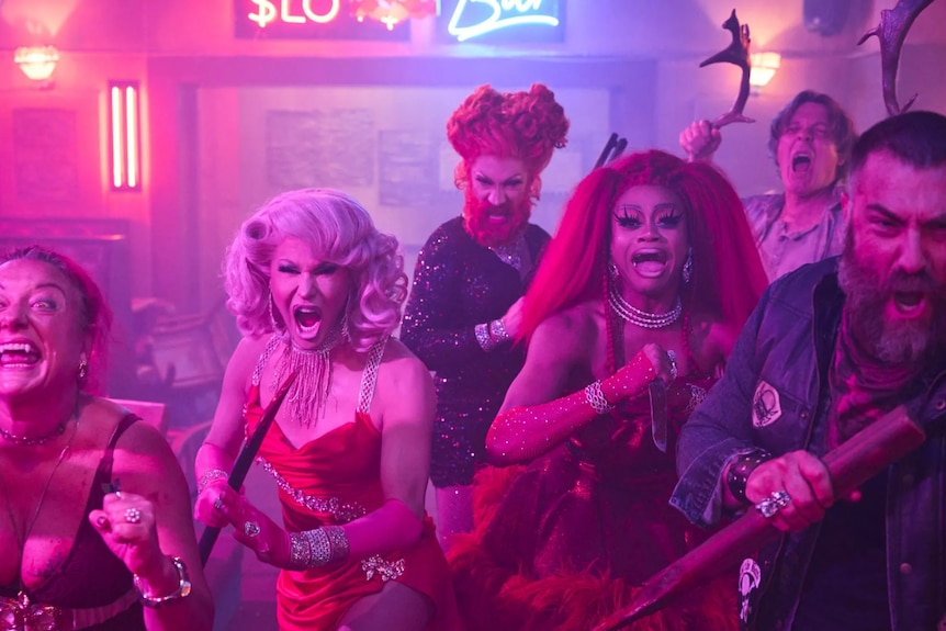 Cinq personnes - trois d'entre elles sont des drag queens - et un vampire brandissent des armes, prêts à se battre.