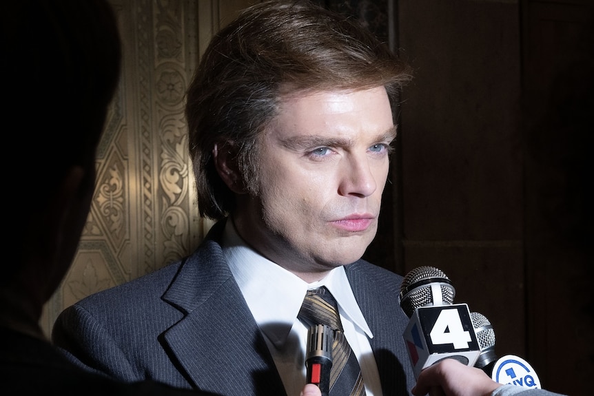 Un homme en costume bleu grince des dents tandis que des journalistes tiennent des micros devant son visage.