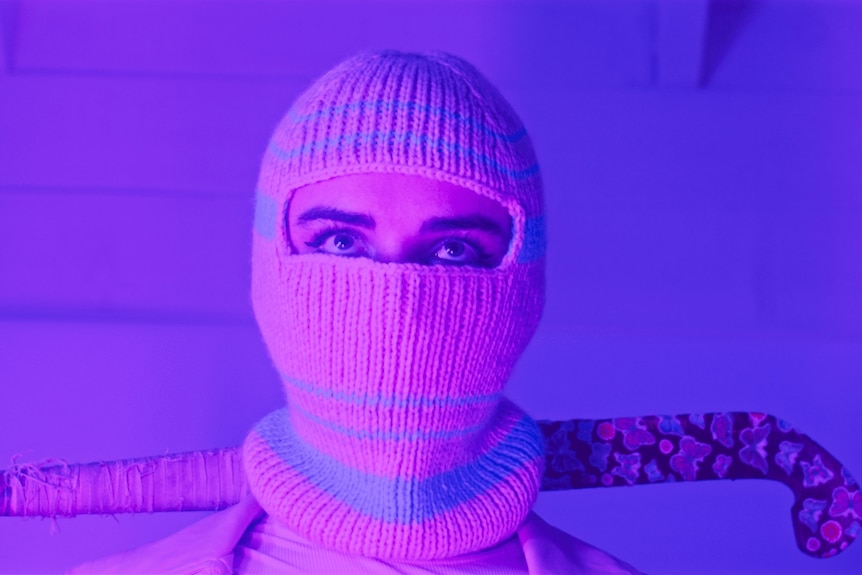 Une femme en cagoule tient une crosse de hockey sous un éclairage rose et violet.