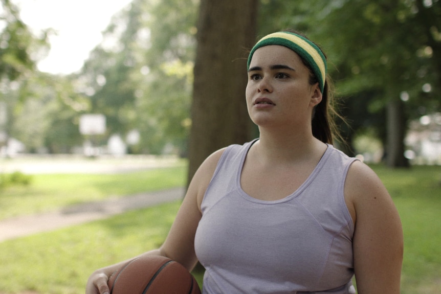 Une femme portant un débardeur et un bandeau tient un ballon de basket sous son bras droit dans un parc.
