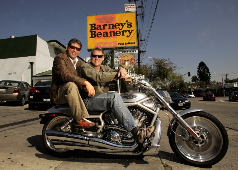 Two men on a motorcycle pose in front of a sign that reads "Barney's Beanery."