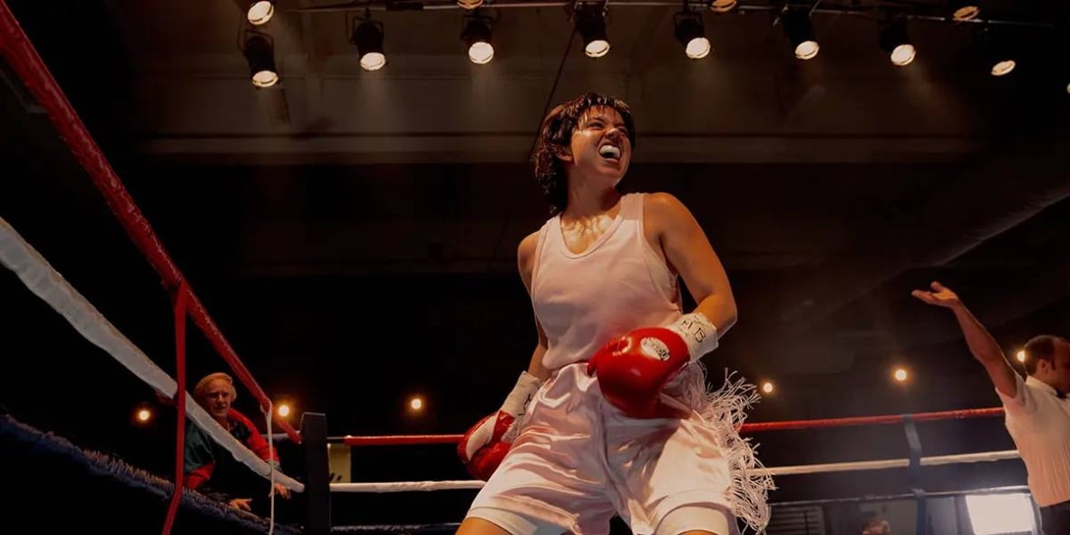 Une femme se tient dans un ring de boxe sous des lumières brillantes. Elle porte un équipement de boxe et sourit largement, révélant un protège-dent.