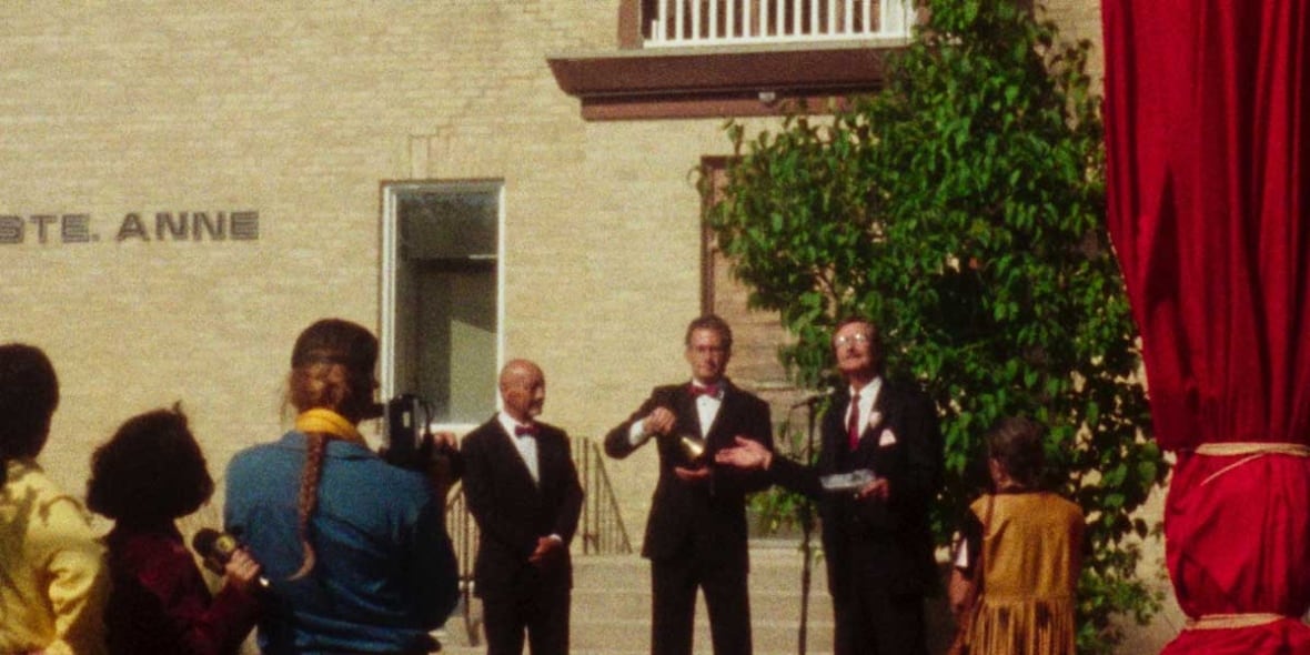 Une image granuleuse d'une foule regardant un trio d'hommes en costumes sombres. Le trio se tient devant des microphones. Une église en briques se trouve derrière eux.
