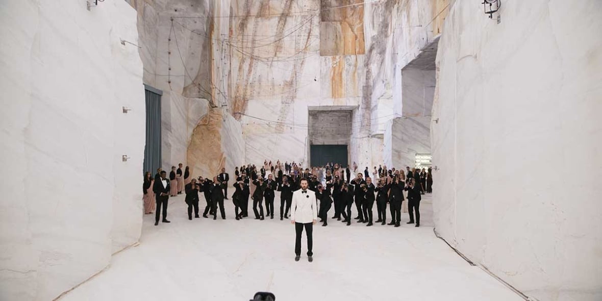 Vue large d'une pièce blanche à plafond haut. Un homme en smoking blanc se tient devant une foule habillée en noir. Beaucoup semblent prendre des photographies de l'homme en smoking.