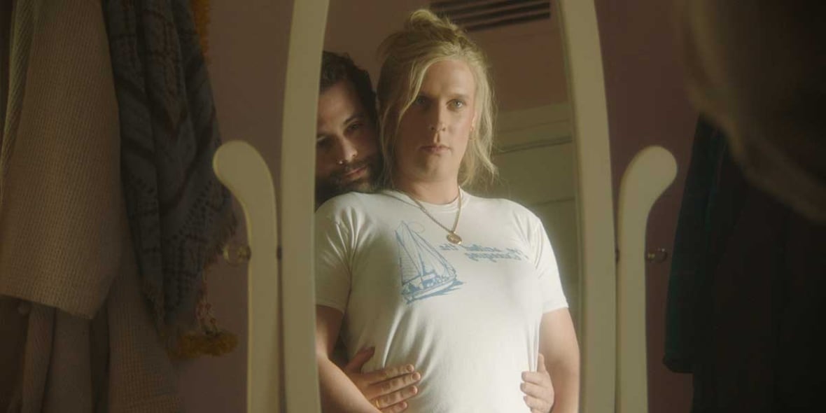Deux personnes dans une faible lumière se tiennent devant un miroir. La personne devant porte un t-shirt blanc avec un design graphique. La personne derrière tient la taille de son compagnon tout en reposant sa tête sur l’épaule de l’autre.
