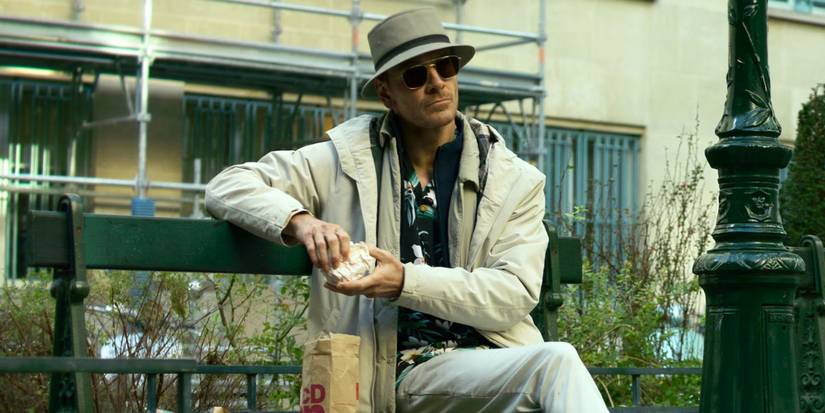Michael Fassbender assis sur un banc, portant des lunettes de soleil et une tenue beige, dans The Killer