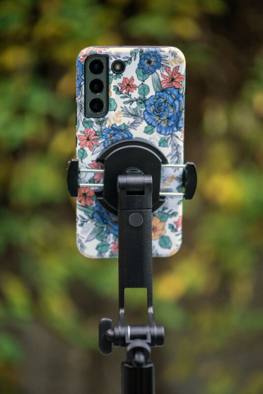 Un smartphone avec une coque fleurie est monté sur un trépied noir, avec un fond flou de feuillage vert et jaune.