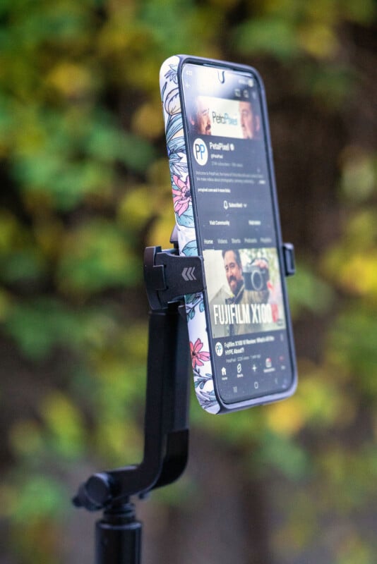 Un smartphone avec une coque à motifs floraux est monté sur un trépied, affichant une page de chaîne YouTube. L'arrière-plan est un flou de verdure.