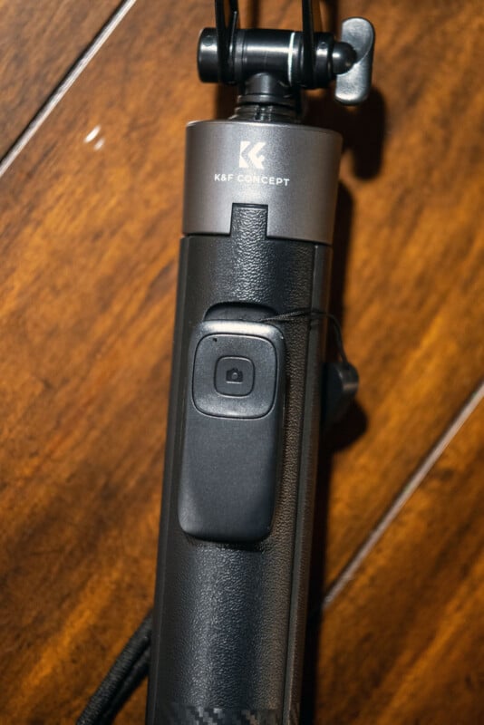 Gros plan d'une poignée noire de trépied ou de perche K&F Concept avec un bouton de télécommande intégré, reposant sur une surface en bois.