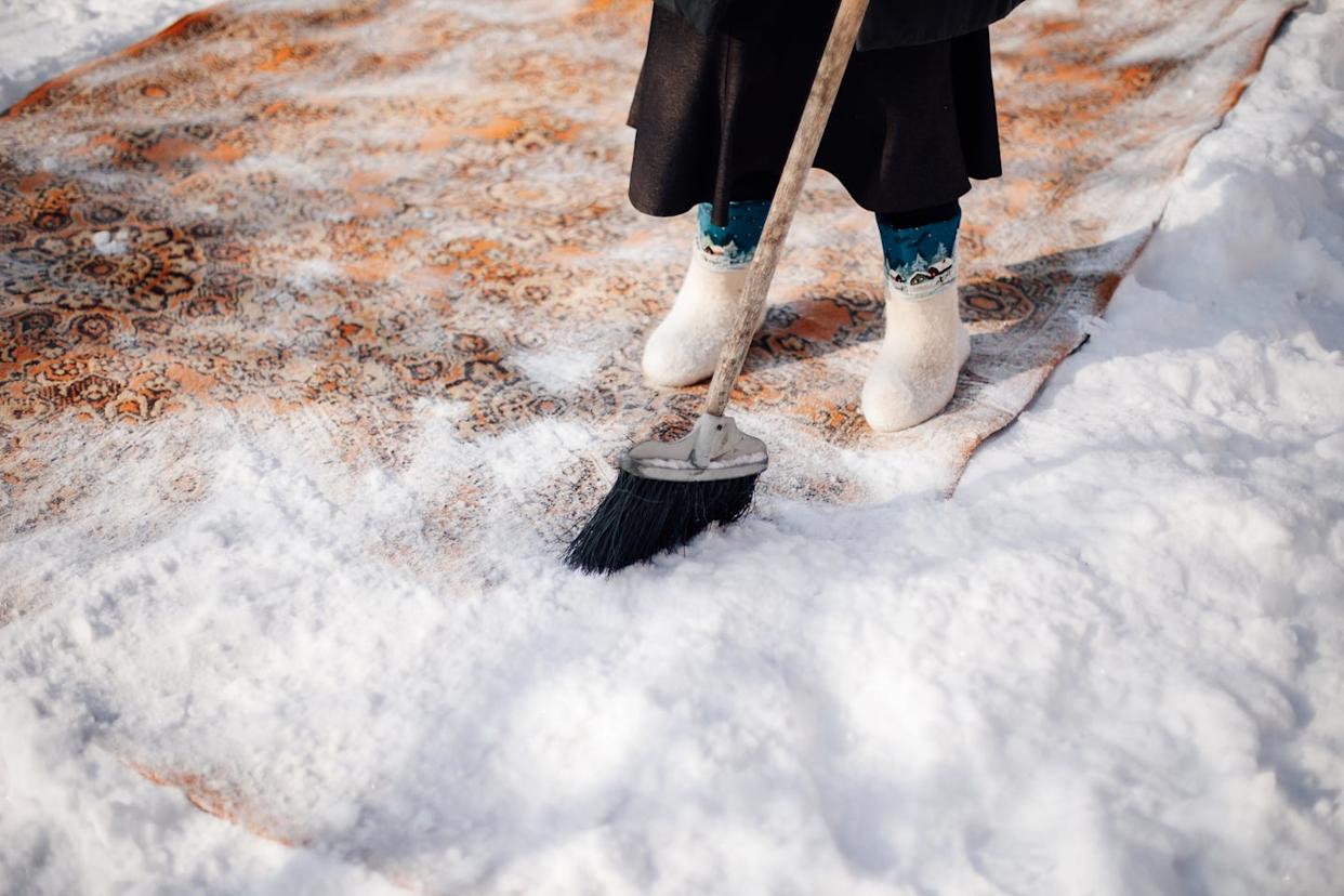Un homme en bottes en feutre se tenant sur un tapis couvert de neige, utilisant un balai pour enlever la neige