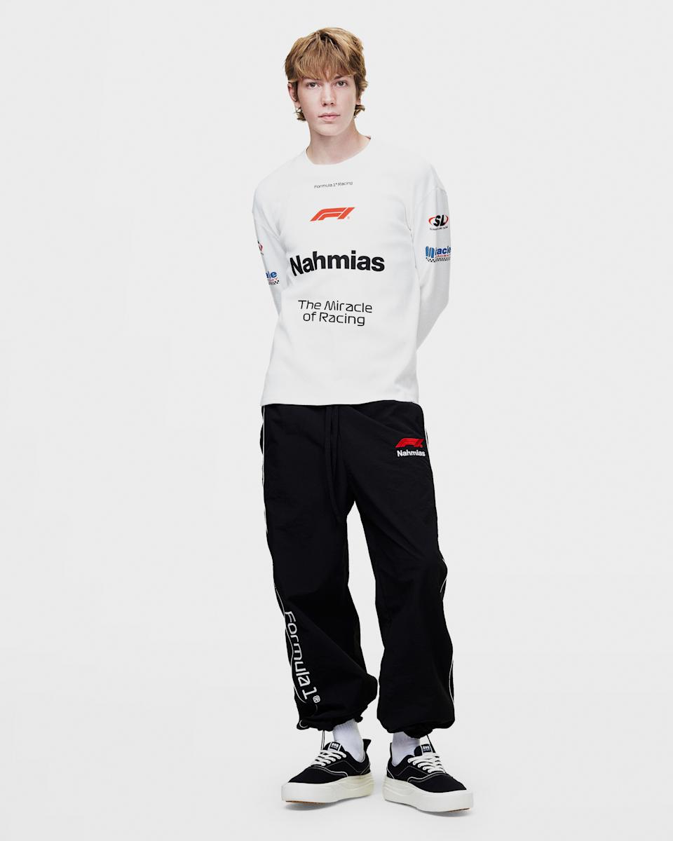 Une personne avec des cheveux châtain clair portant un haut à manches longues blanc et un pantalon noir avec les marques 'Nahmias' et 'Formula 1', se tenant contre un fond uni.