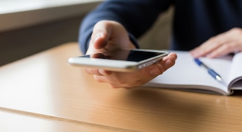Un adolescent tenant un téléphone mobile sur un bureau en bois clair, avec un carnet et un stylo.