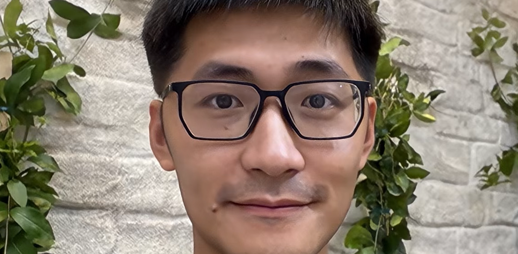 Autoportrait d’un homme avec des lunettes devant un mur de pierre blanche avec des plantes.