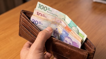 Une personne tient un portefeuille en cuir marron contenant des billets colombiens sur une table en bois clair.
