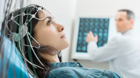 Femme allongée sur un lit médical avec des électrodes. En arrière-plan, un professionnel de santé examine des images de scanner cérébral.