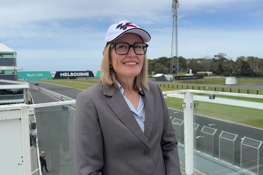 Une femme blonde portant des lunettes et une casquette MotoGP se tenant près de la piste.