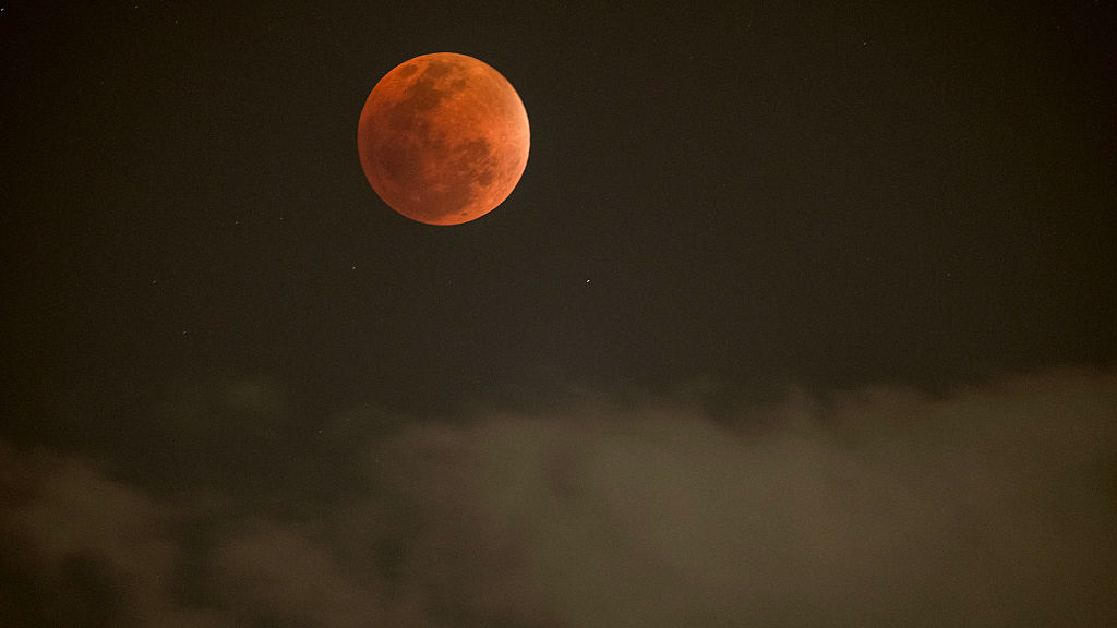 Une pleine lune rouge est photographiée au-dessus de nuages gris dans un ciel nocturne sombre.