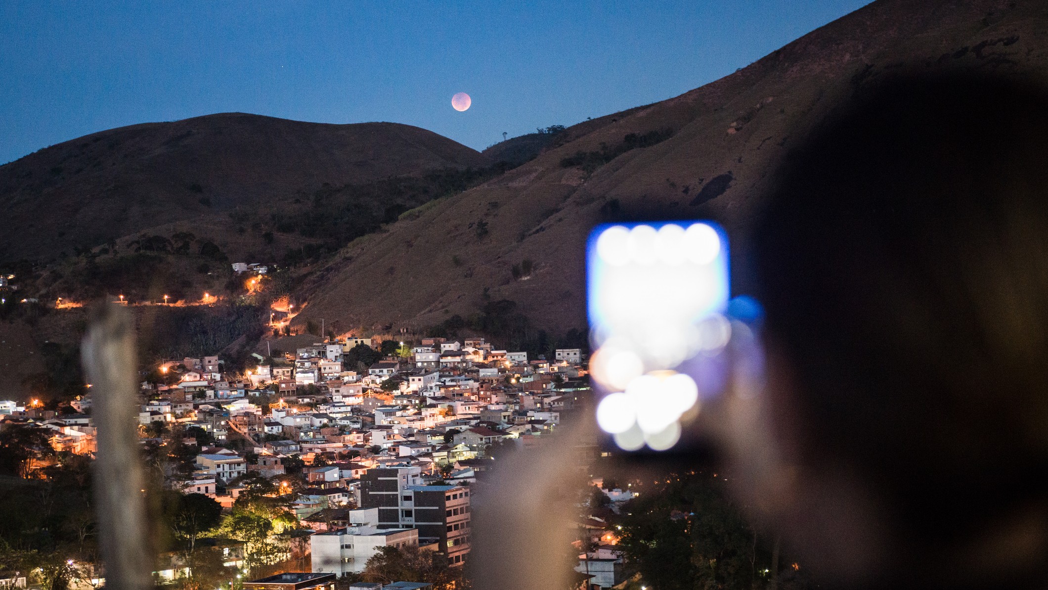 une personne tenant un téléphone pour prendre une photo d'une éclipse lunaire totale se produisant au-dessus d'une colline avec des lumières de ville en dessous.