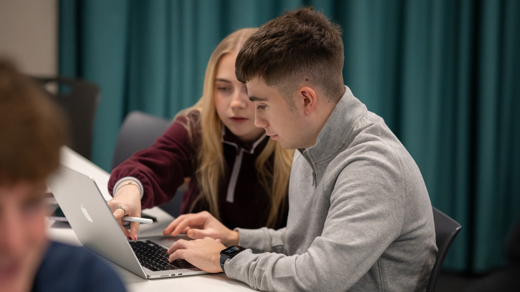 Des camarades de classe collaborent sur un MacBook, l'une pointe l'écran pendant que l'autre tape.