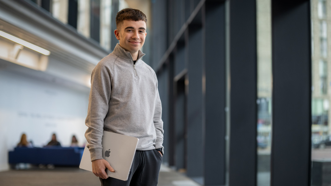 Un étudiant se tient dans un couloir moderne avec un ordinateur portable MacBook, souriant à la caméra
